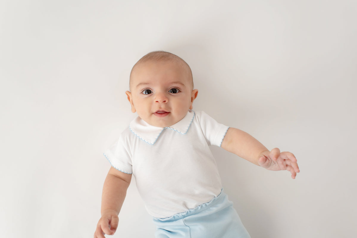 Bebê de body branco com gola e calça azul clara deitado em fundo claro passalinho
