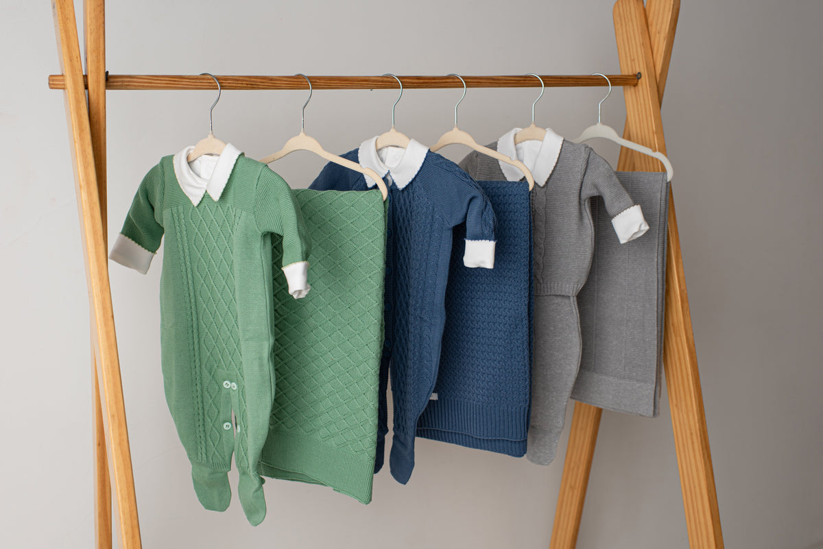 Saídas de maternidade em tricô verde, marinho, azul e cinza penduradas em arara de madeira passalinho