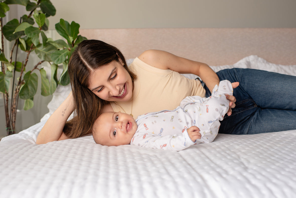 Mãe deitada na cama sorrindo para o bebê de macacão estampado passalinho