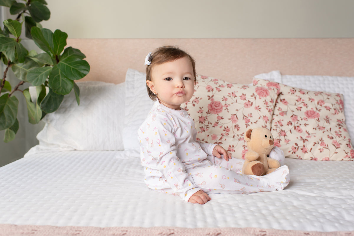 Bebê de macacão branco sentada na cama com almofadas floridas e ursinho passalinho